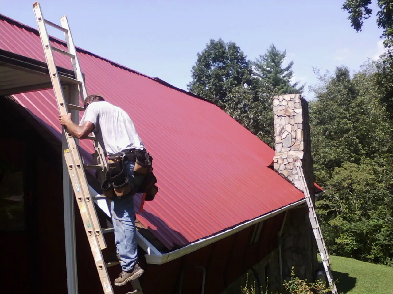 Roofer on a ladder beside a red metal roof and stone chimney — Soffit & Fascia Repair by Roofing Brothers in Solomon